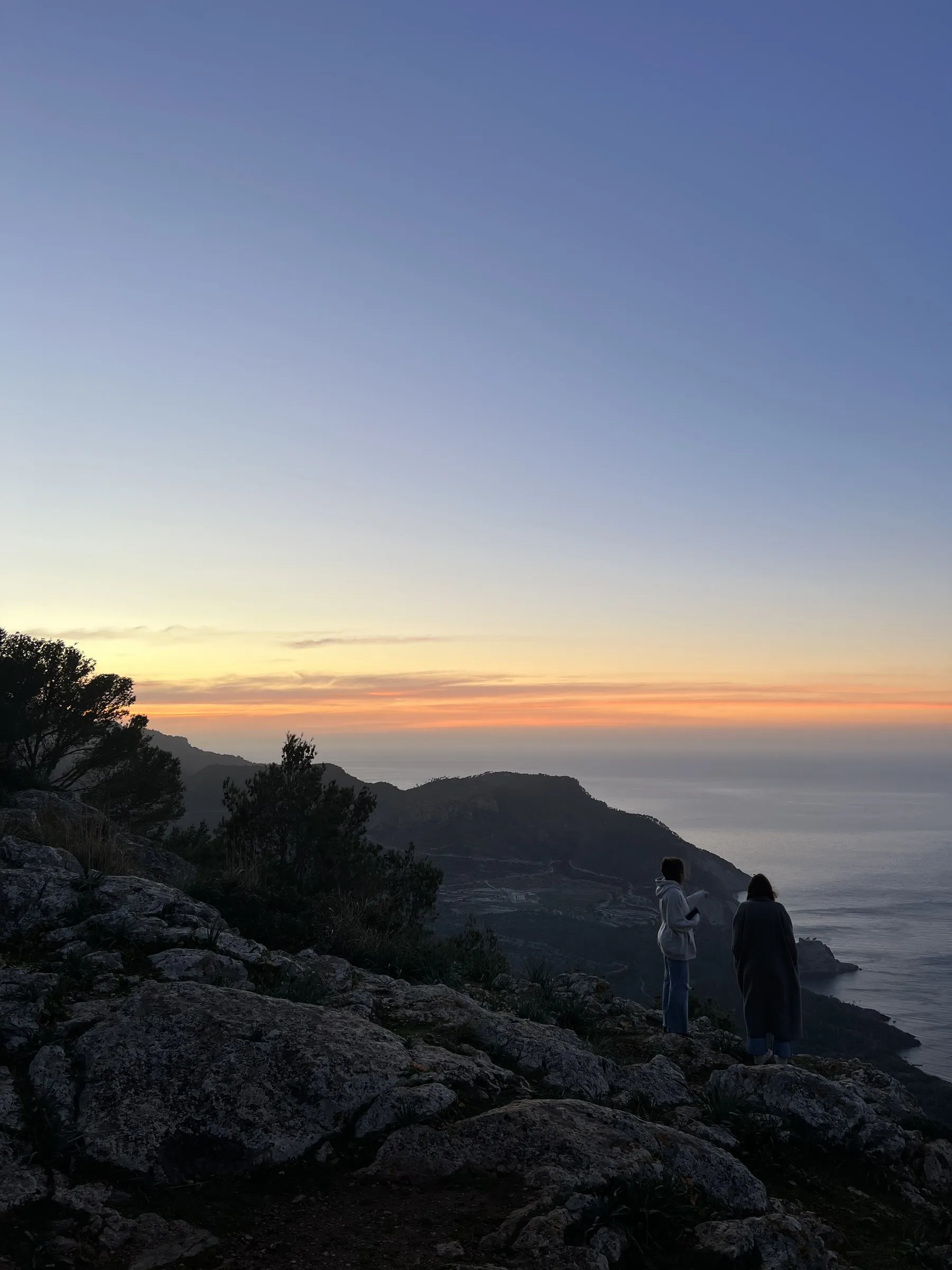 Dos personas de pie sobre las rocas mirando el mar de Banyalbufar mientras se apaga la luz.