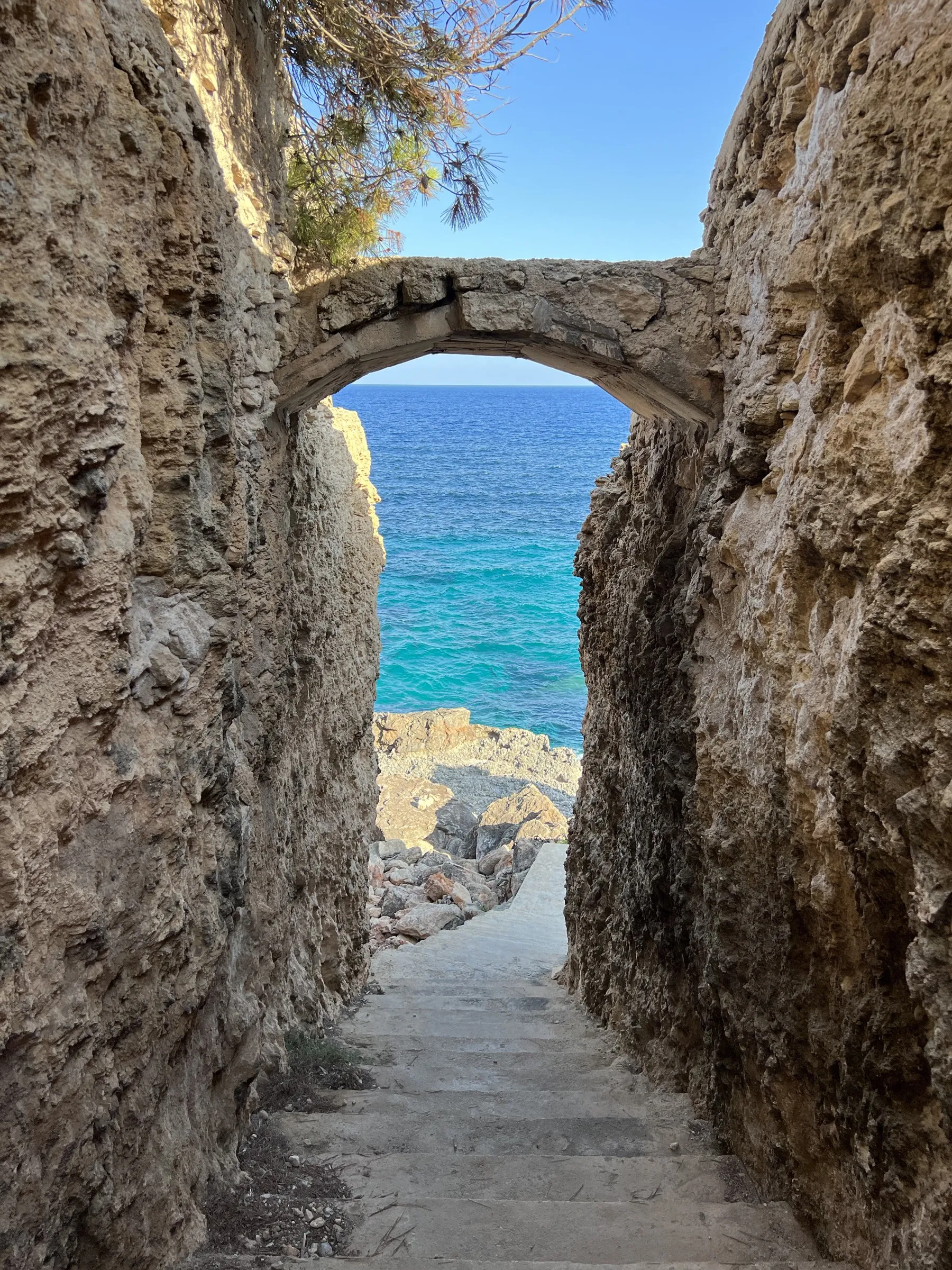 Ein in den Fels gehauener Steinbogen, dahinter Stufen, die zum türkisfarbenen Wasser einer kleinen Bucht hinabführen.