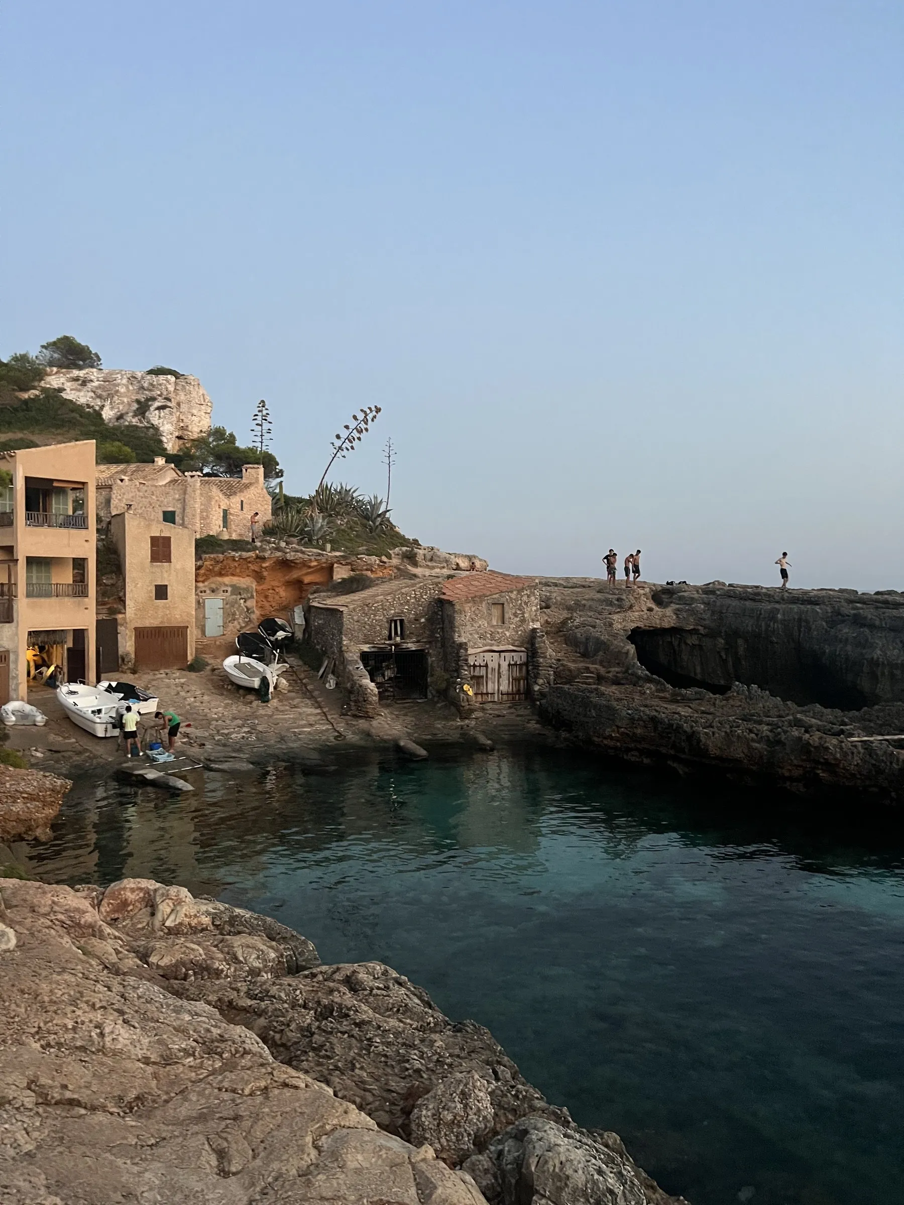 Los escars de piedra de Cala s'Almunia al borde del agua, con barcas varadas y gente sentada en las rocas.