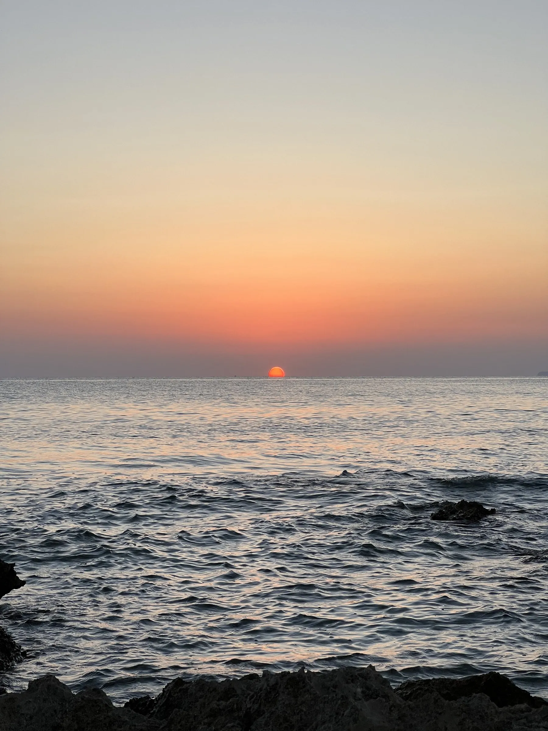 El sol tocando el horizonte sobre el mar abierto frente al Cap de ses Salines, con las rocas bajas en primer término.